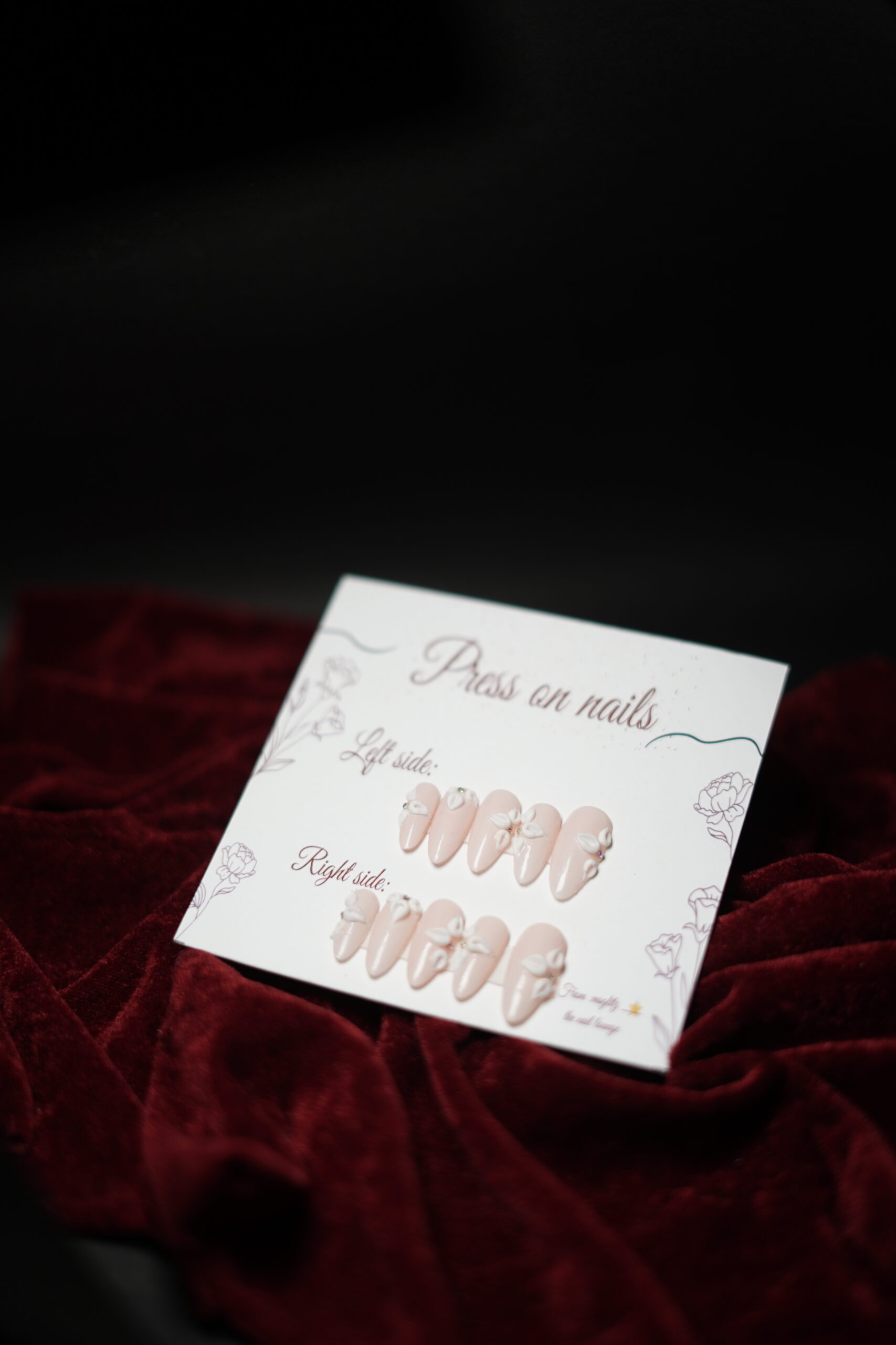 Set of nude press-on nails with 3D white floral accents displayed on a card labeled left side and right side, placed on red velvet fabric.”