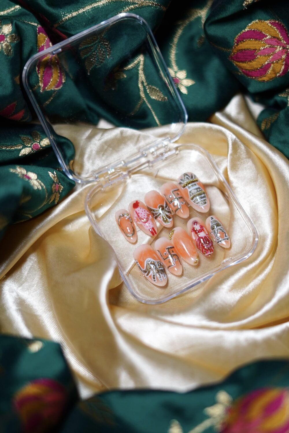 A set of peach-toned press-on bridal nails with intricate hand-painted designs including a bride in red lehenga, doli, mandap, kalash, floral details, and glitter embellishments. The middle nail in the second row is left blank for customized initials of the bride and groom.