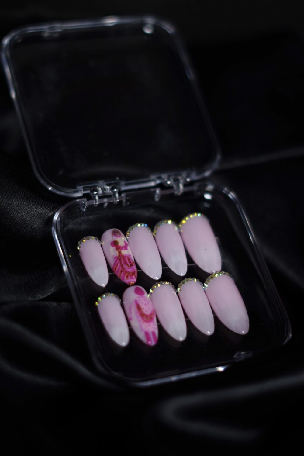 A set of pink ombré press-on nails with glossy finish, decorated with rhinestone tips and intricate mehendi-inspired red detailing on accent nails, placed inside a clear box on a black satin background.